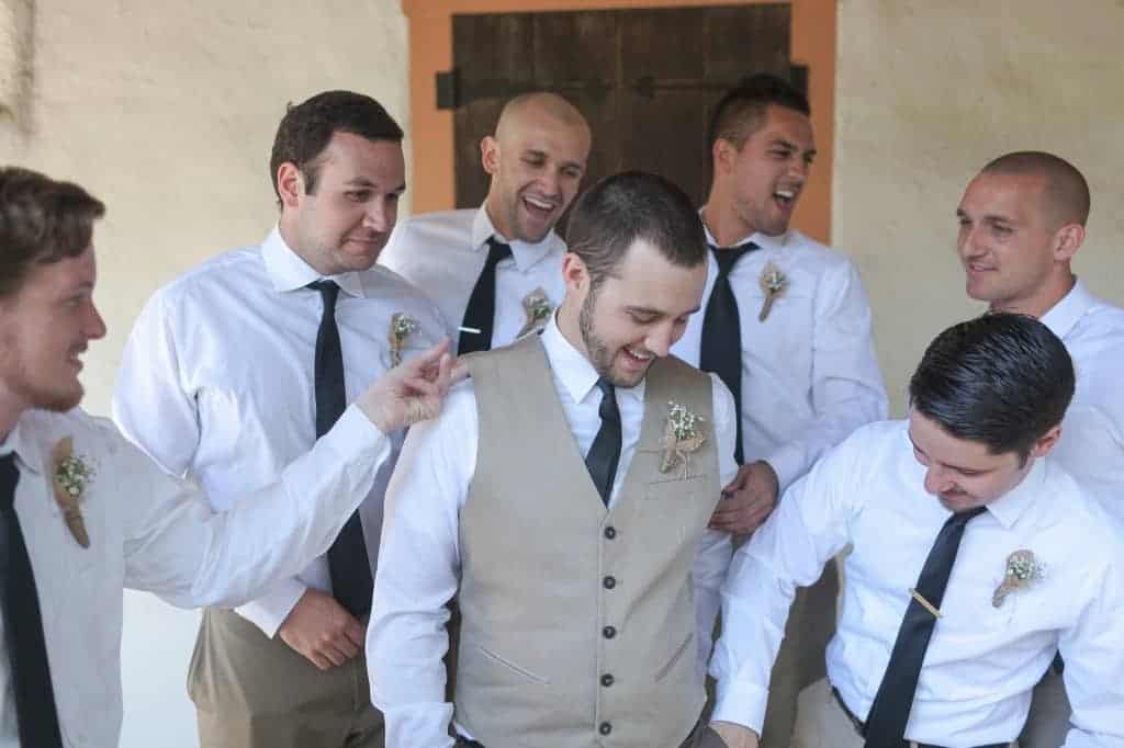 The groom and his wedding party. The groomsmen are wearing white shirts, black ties, and khaki dress pants. The groom is wearing a white shirt, black tie, and a khaki colored vest.