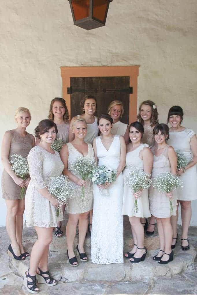 Bridesmaids standing around bride wearing neutral colored bridesmaid dresses holding baby's breath bouquets with door behind them