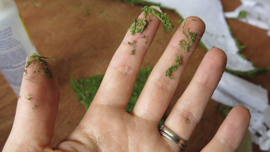 A picture of a woman's hands with faux crafting moss stuck all over her fingers.