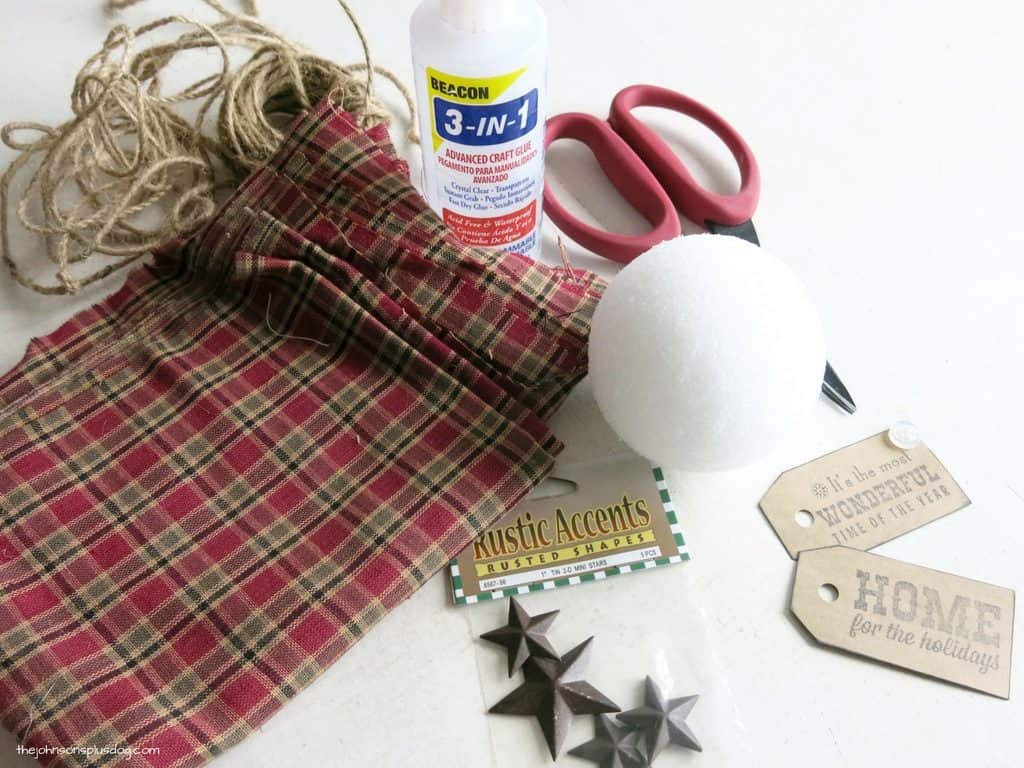 The supplies needed to make the second kind of homemade fabric ornament - a sheet of red plaid homespun fabric, jute twine, craft glue, scissors, a styrofoam craft ball, and rustic decor accents.