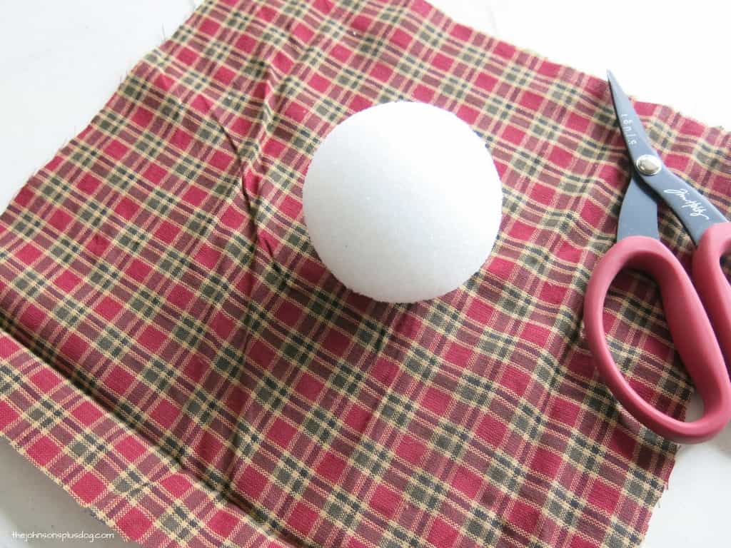 A large square of red and green plaid homespun fabric with a styrofoam craft ball and craft scissors sitting on a white table.