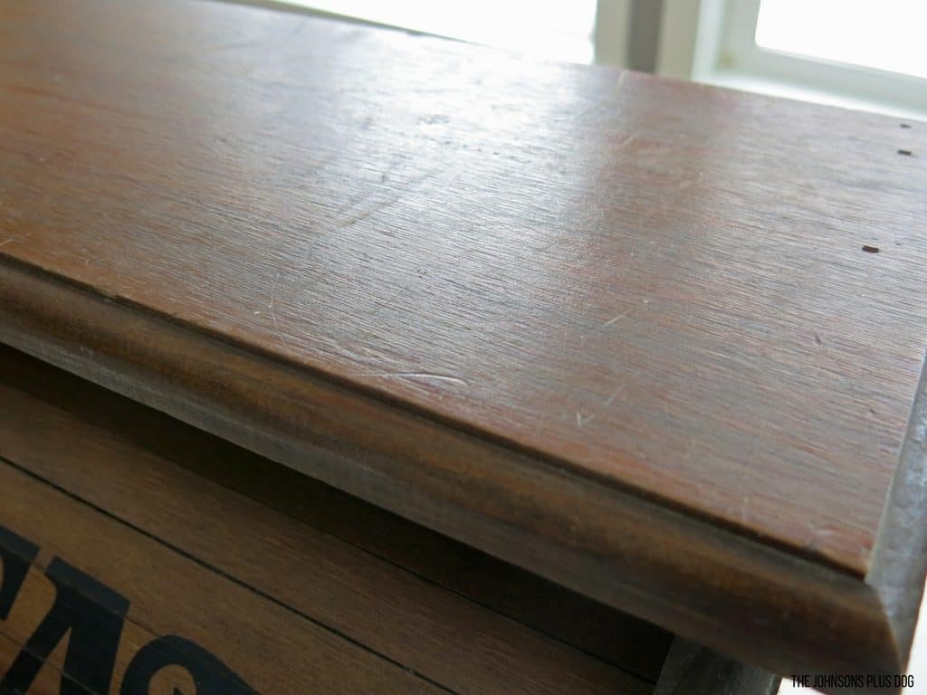 A close up look at the top of the vintage bread box, showing scratches and dents in the stained wood surface.