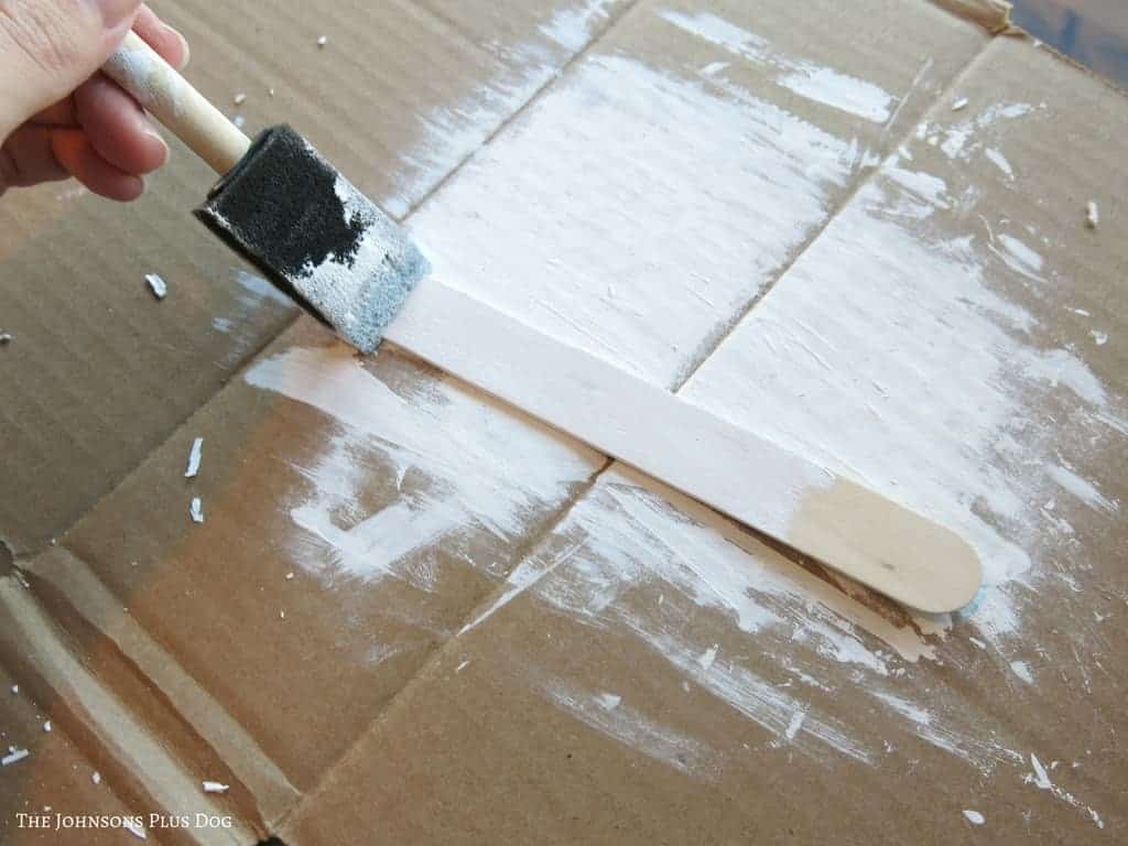 hand holding foam paint brush painting a popsicle stick lying on cardboard