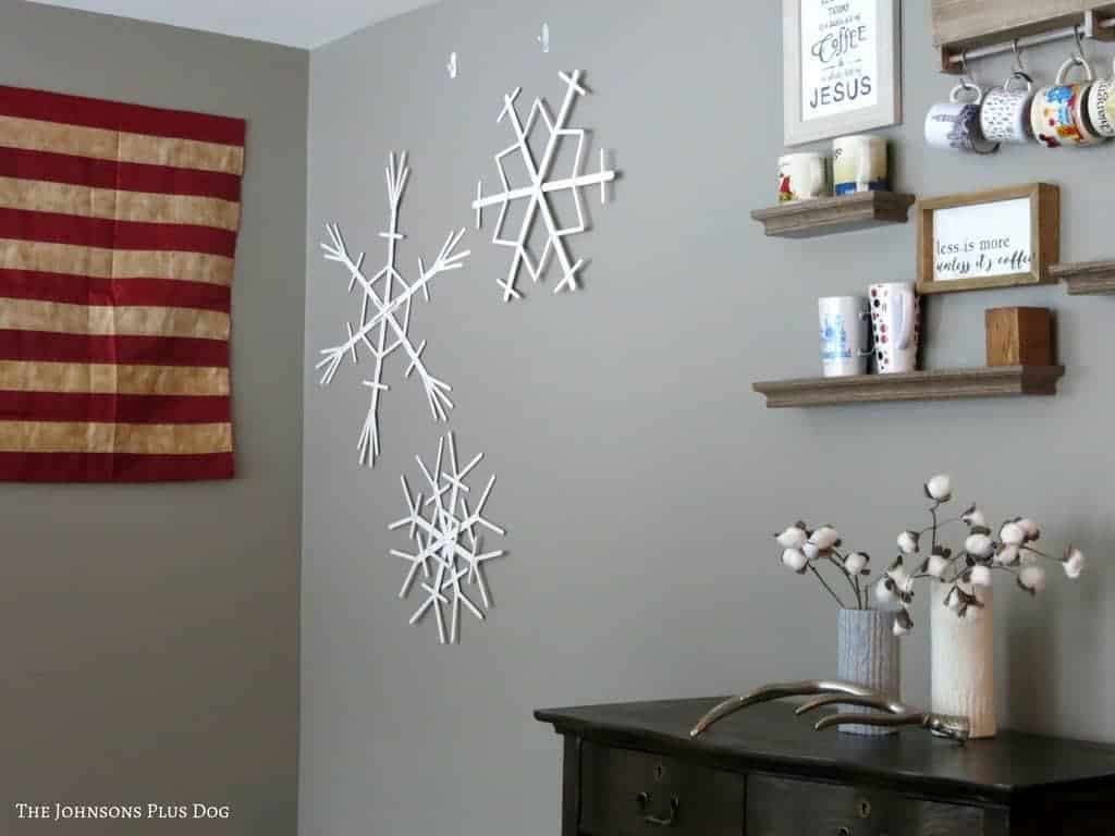 Popsicle sticks snowflakes on the wall beside the wall frame, hanging shelves with mugs vases with cotton stern on top of a farmhouse drawer