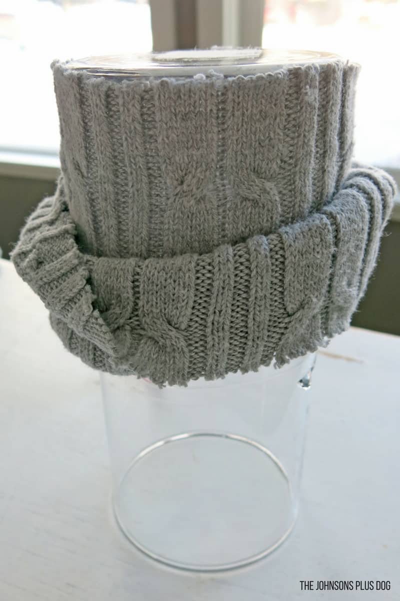 The detached sleeve of a gray cable knit sweater being rolled down a glass vase, to create a cozy sweater covered vase for winter decor.