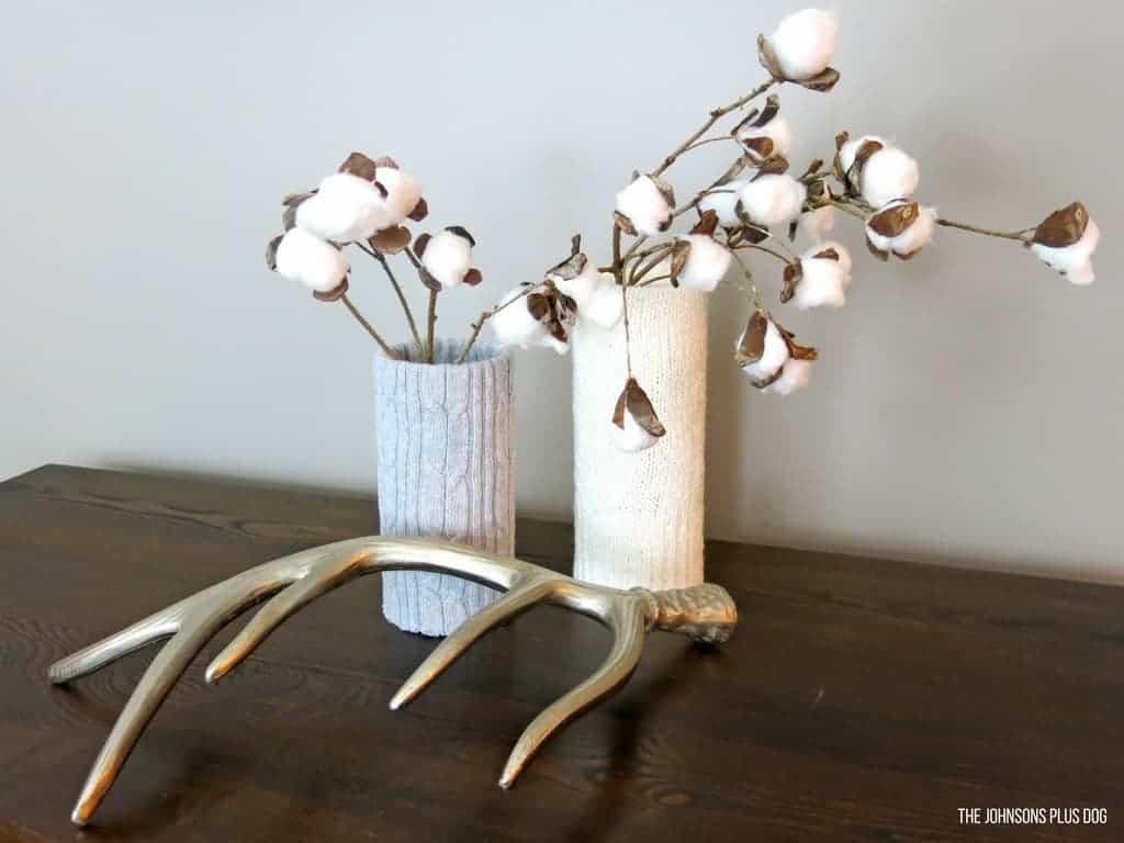 shows two vases covered in sweater material, one gray and the other white on a dark wood table with faux cotton stems in them and next to a metal deer antler