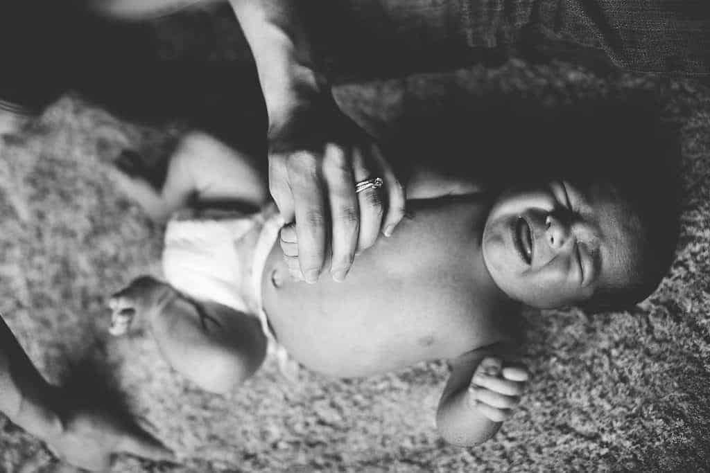 Black and white image of newborn baby crying on the floor wearing a diaper and mom is holding hand