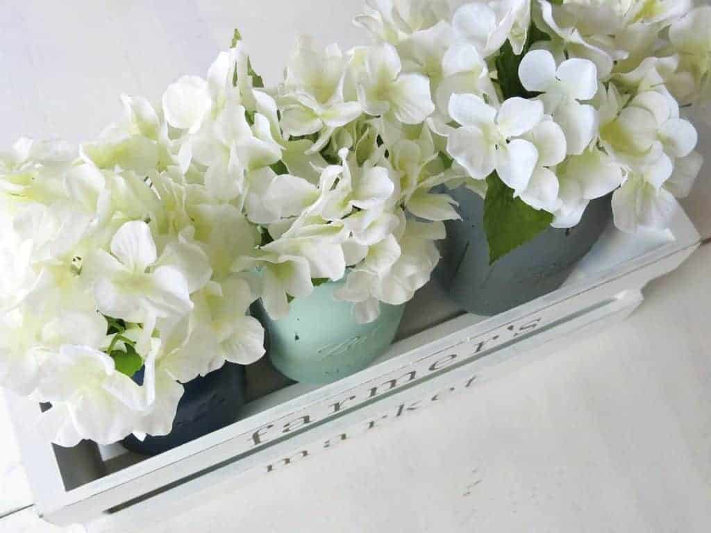 Three painted mason jars lined up with faux white Dollar Store hydrangeas in a white painted wooden crate for a Farmhouse centerpiece