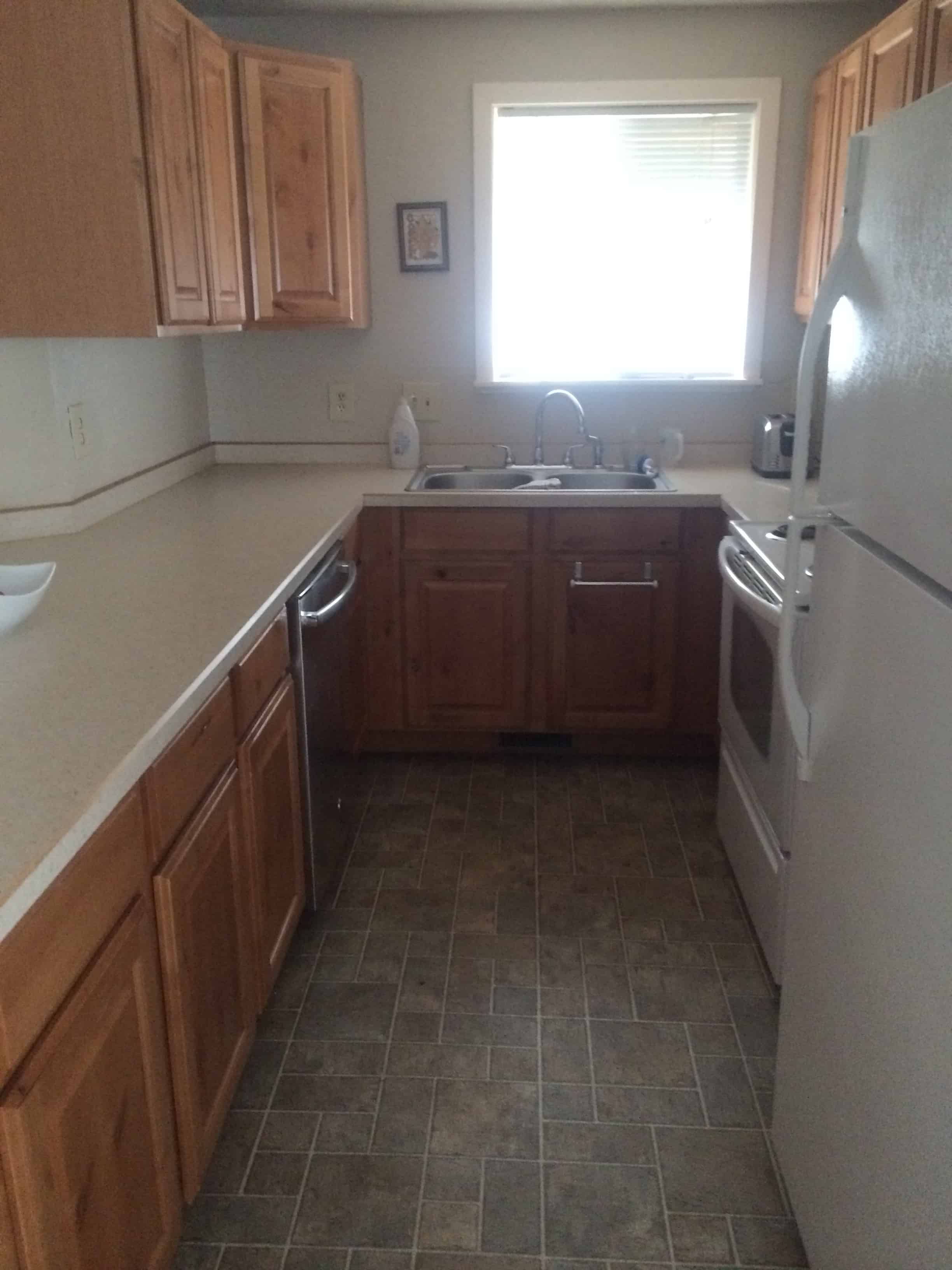 A view inside the kitchen, looking at the small cooking area with countertops, a double sink, oven, and fridge. There are wall-mounted cabinets and a small window.