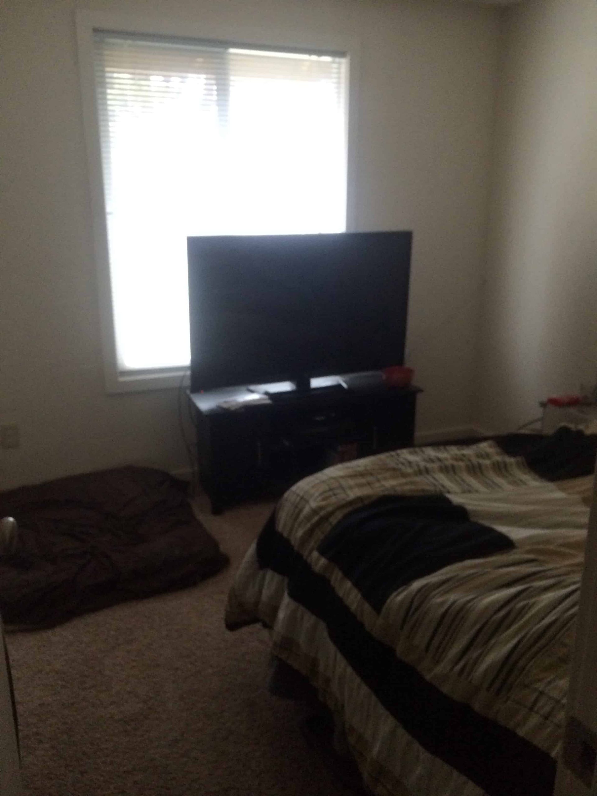The view from the door of our second bedroom shows a large TV on a chest of drawers in front of a bright window, and the foot of a queen sized bed, covered in a quilted comforter. 