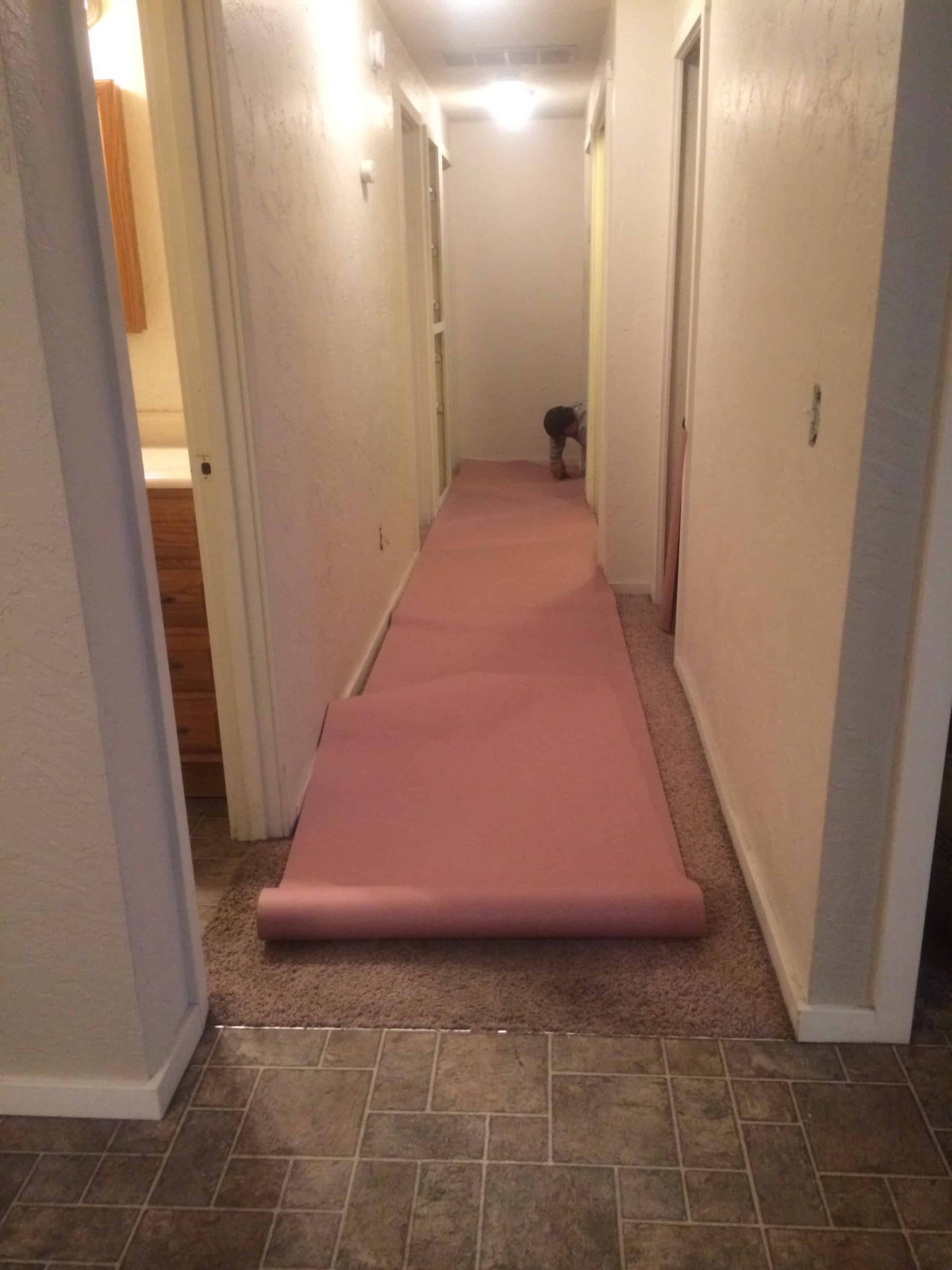 A narrow hallway with white walls and soft lighting. Multiple doors on either side of the hallway are open, leading into branching off rooms. There's a man at the end of the hallway rolling protective covering over the brown carpet.