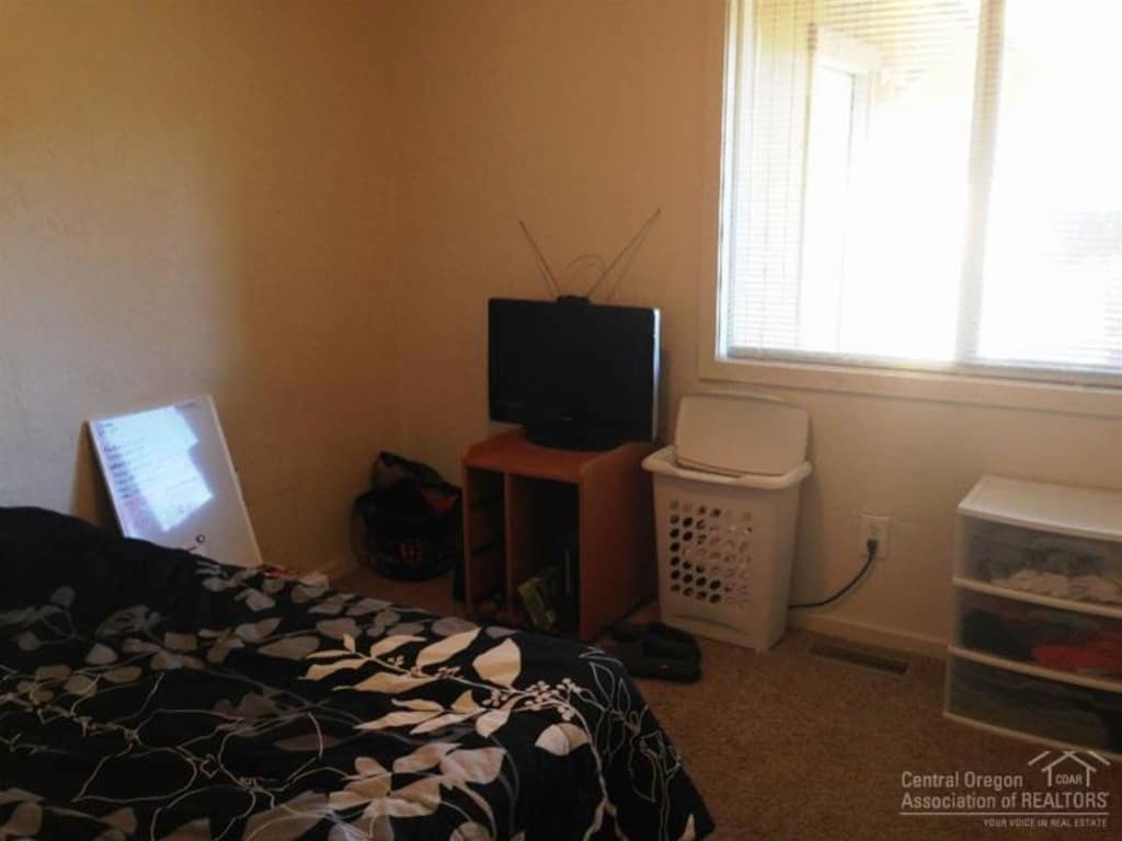 An image of a cluttered corner of a bedroom, where a small TV rests on a makeshift tv stand next to a clothing hamper and stacked plastic drawers, under a large window.
