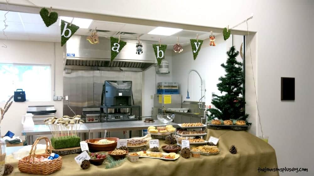 A rustic woodland animal themed baby shower banner hangs on a wall over a buffet filled with trays of food and snacks.