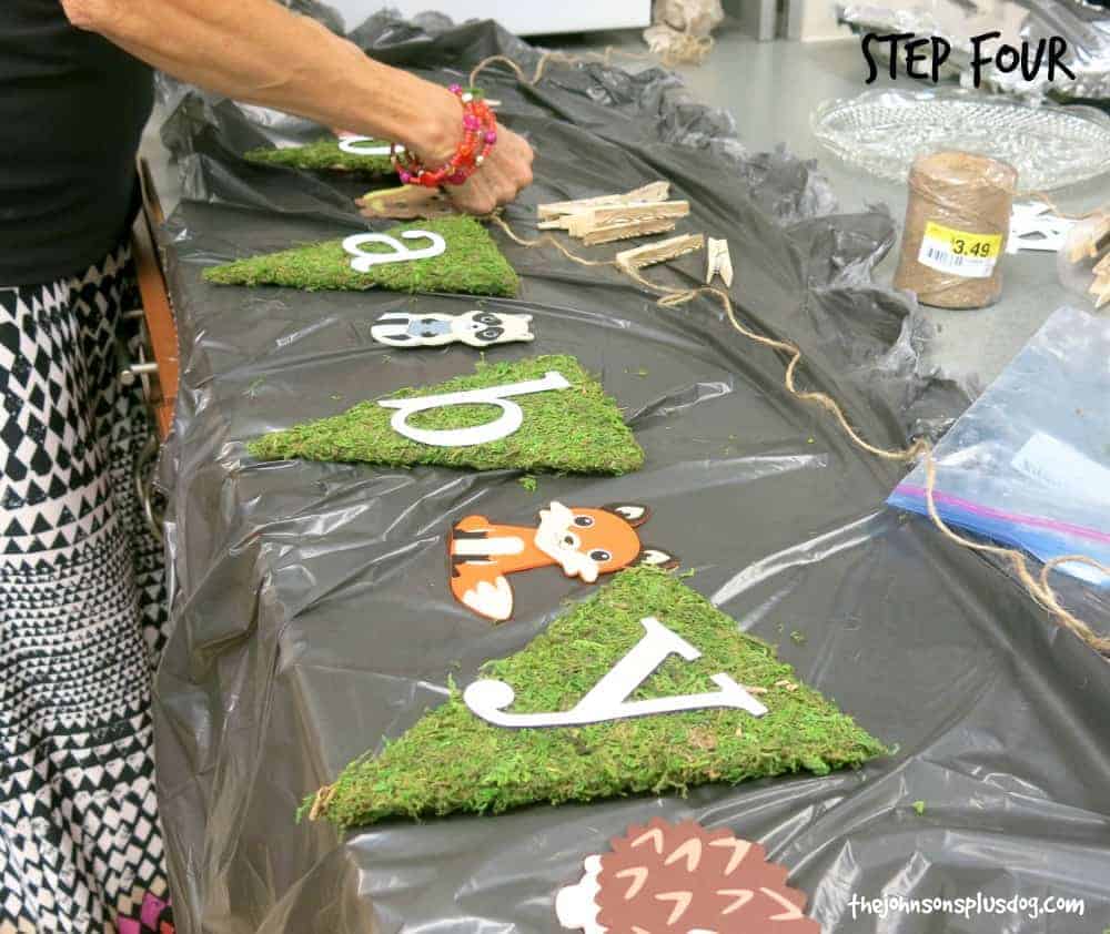 A woman works on stringing together all the elements for a rustic woodland animal themed baby shower banner. The moss-covered flags and woodland animal pictures lay on a covered table, while a person uses twine to string the flags together.