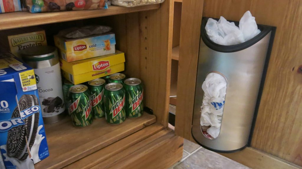 Metal bag holder hanging on door of cabinet turned into DIY pantry. Pantry is stocked with food and drinks like iced team mountain dew and oreos