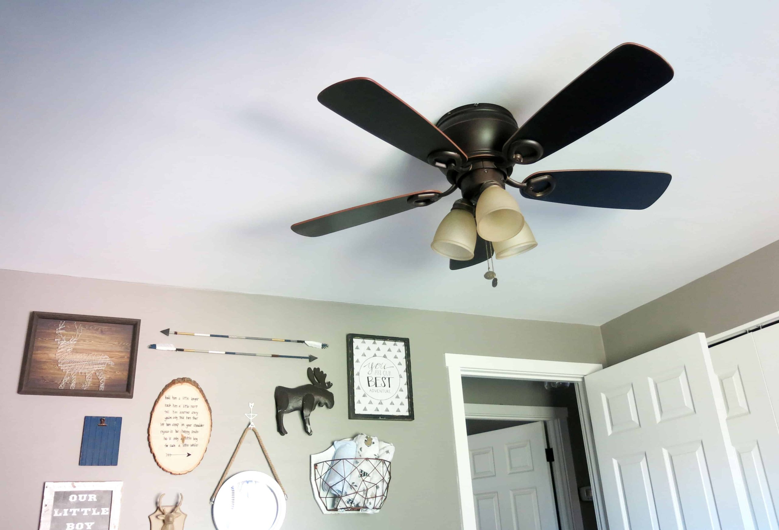 A newly installed ceiling fan on a white ceiling. The fan blades are a deep brown color, and the ceiling fan has three light fixtures. There's a gallery wall filled with rustic style nursery decor.