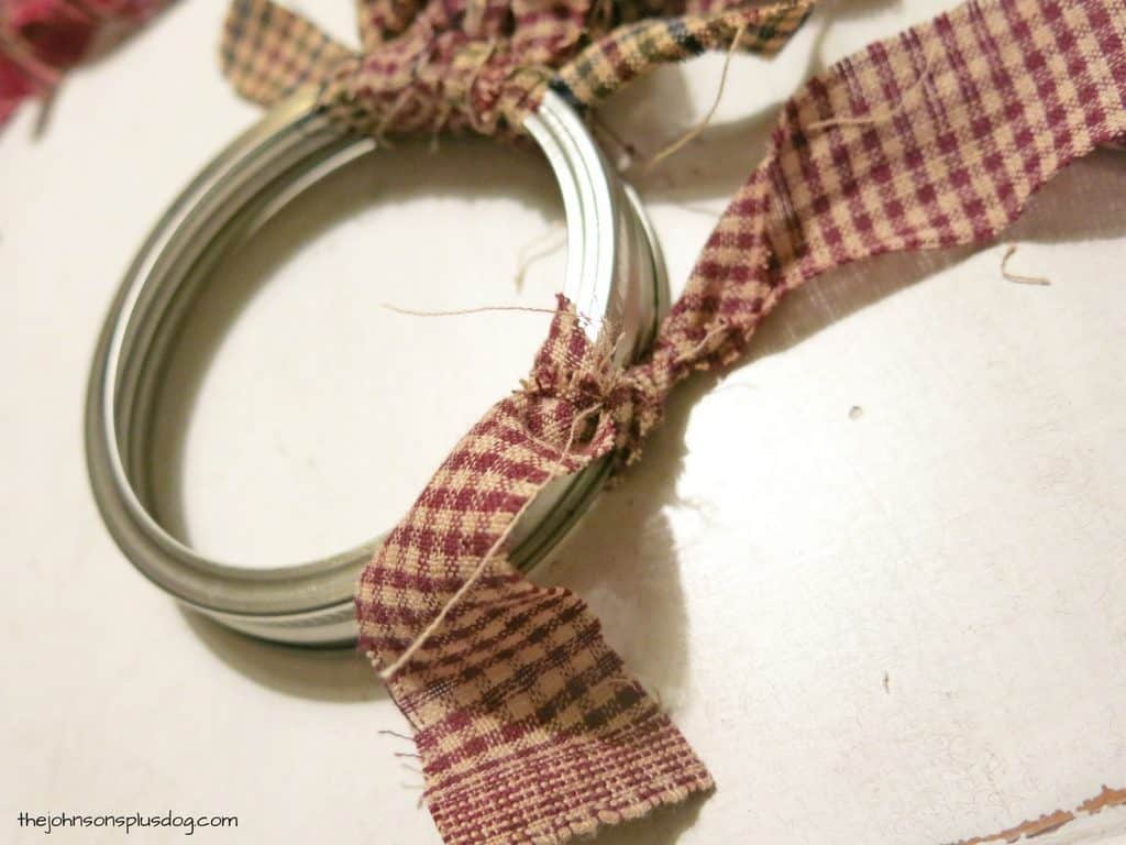 Tying strips of tan and plaid homespun fabric around a mason jar lid to create a homemade fabric ornament.