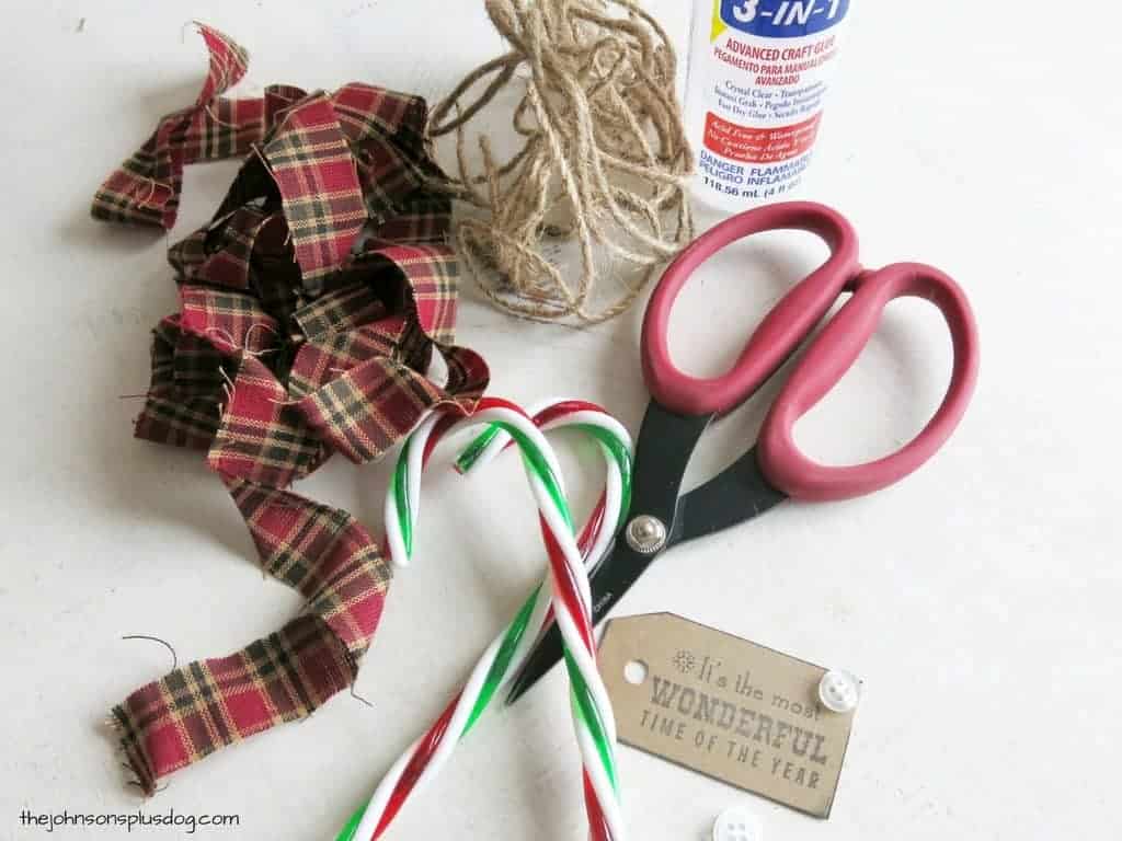 Supplies needed to make candy cane homemade fabric ornaments - two candy canes, strips of red and green plaid fabric, jute twine, scissors, and craft glue.
