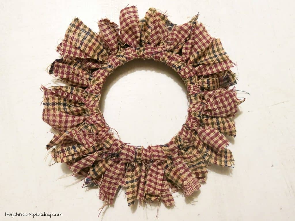 A mini fabric wreath ornament, made with strips of red, green, and tan plaid homespun fabric tied around a mason jar lid.