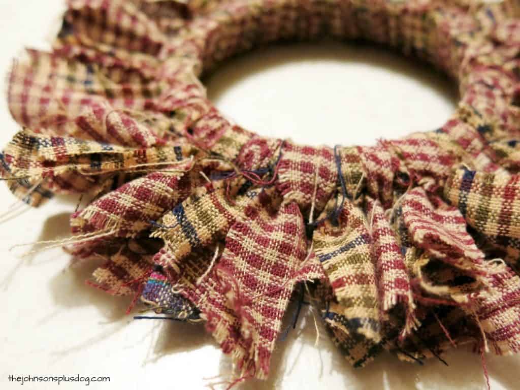 Strips of red, green, and plaid homespun fabric tied with knots around a mason jar lid to create a mini wreath homemade fabric ornament.