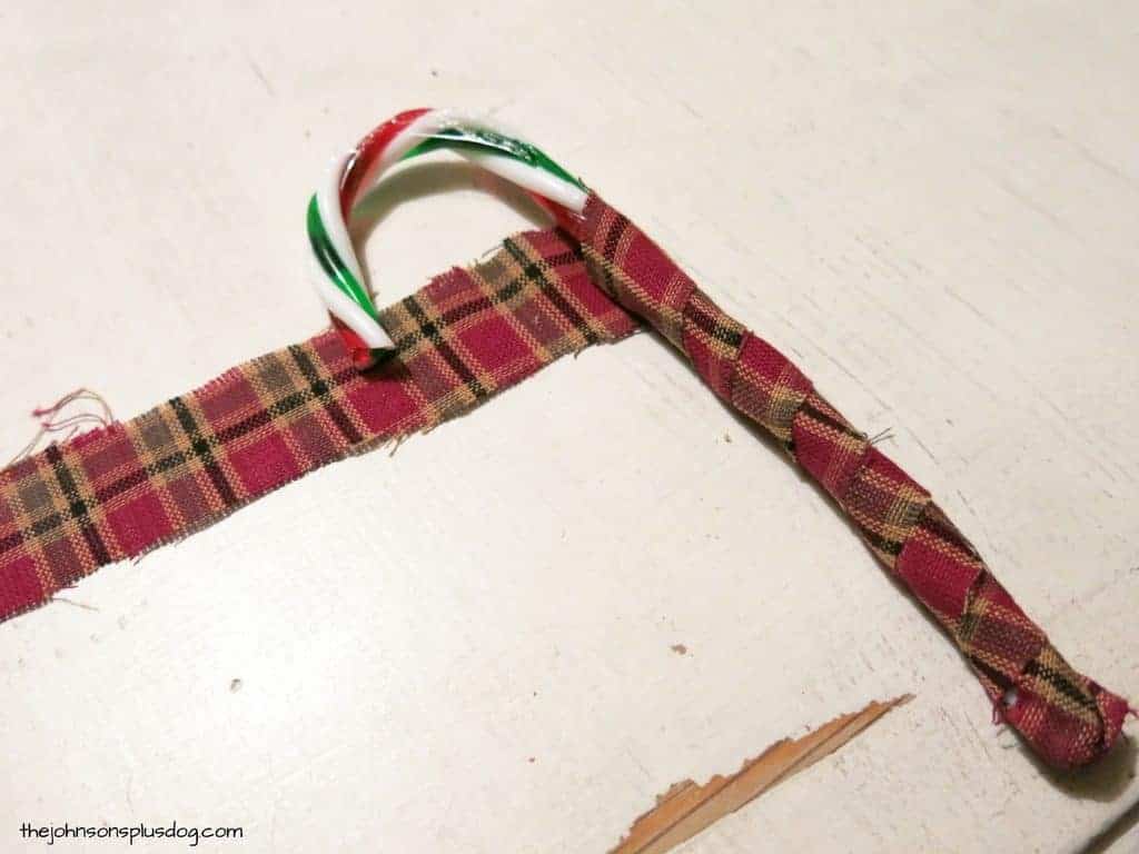 Wrapping a strip of red and tan plaid homespun fabric around a plastic candy cane to create homemade fabric ornaments.