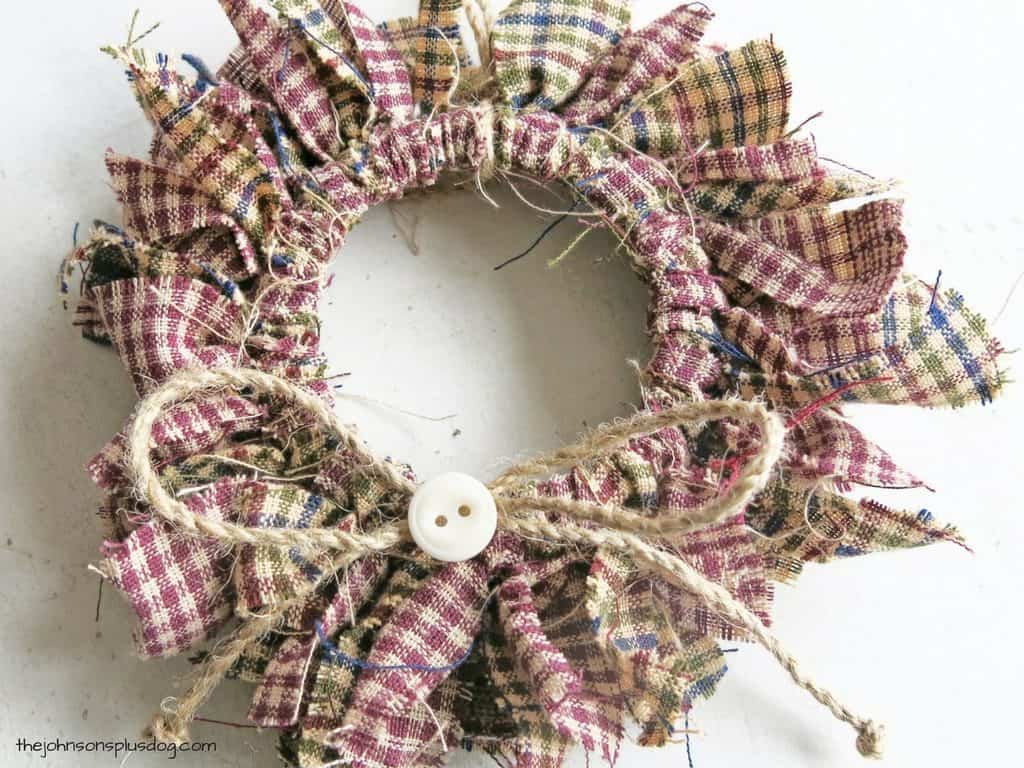 A look at another completed homemade fabric ornament - a mini fabric wreath made with strips of red, green, and tan homespun fabric tied around a mason jar lid