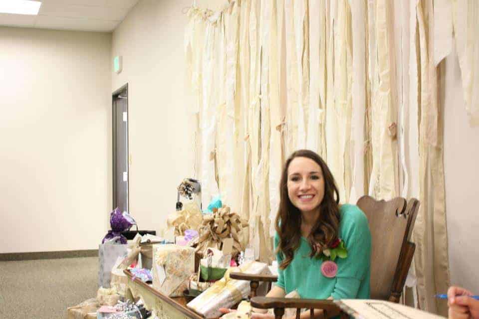 A woman with long brown hair and a green shirt sits in a chair, surrounded by presents, smiling at the camera while attending her bridal shower.