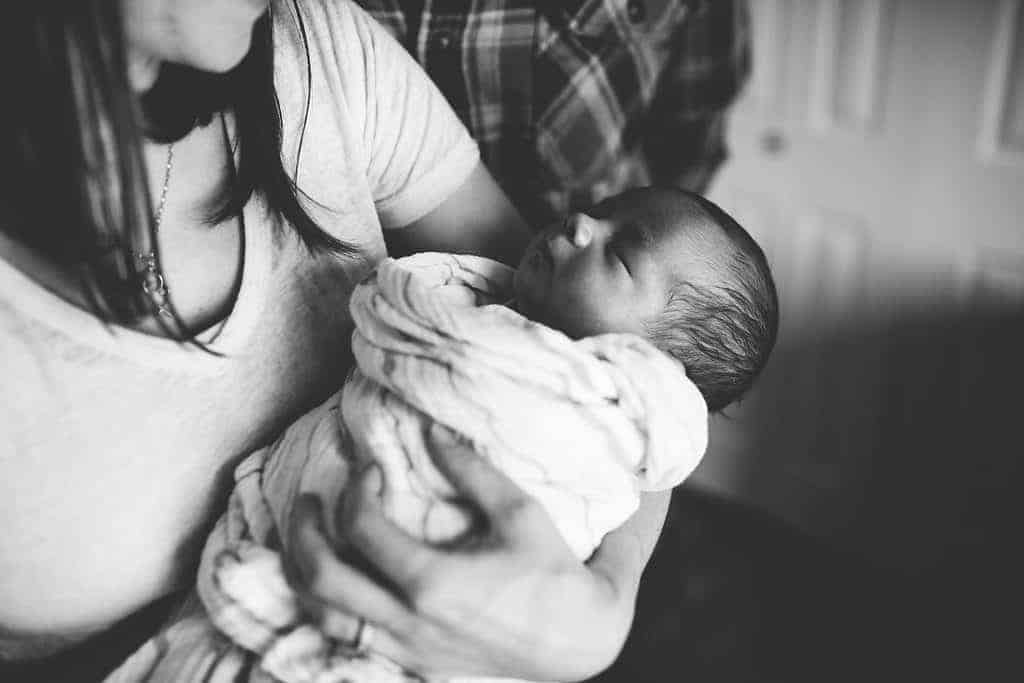 Newborn pictures in black and white with baby wrapped in swaddle blanket behind held by mom and dad standing behind.