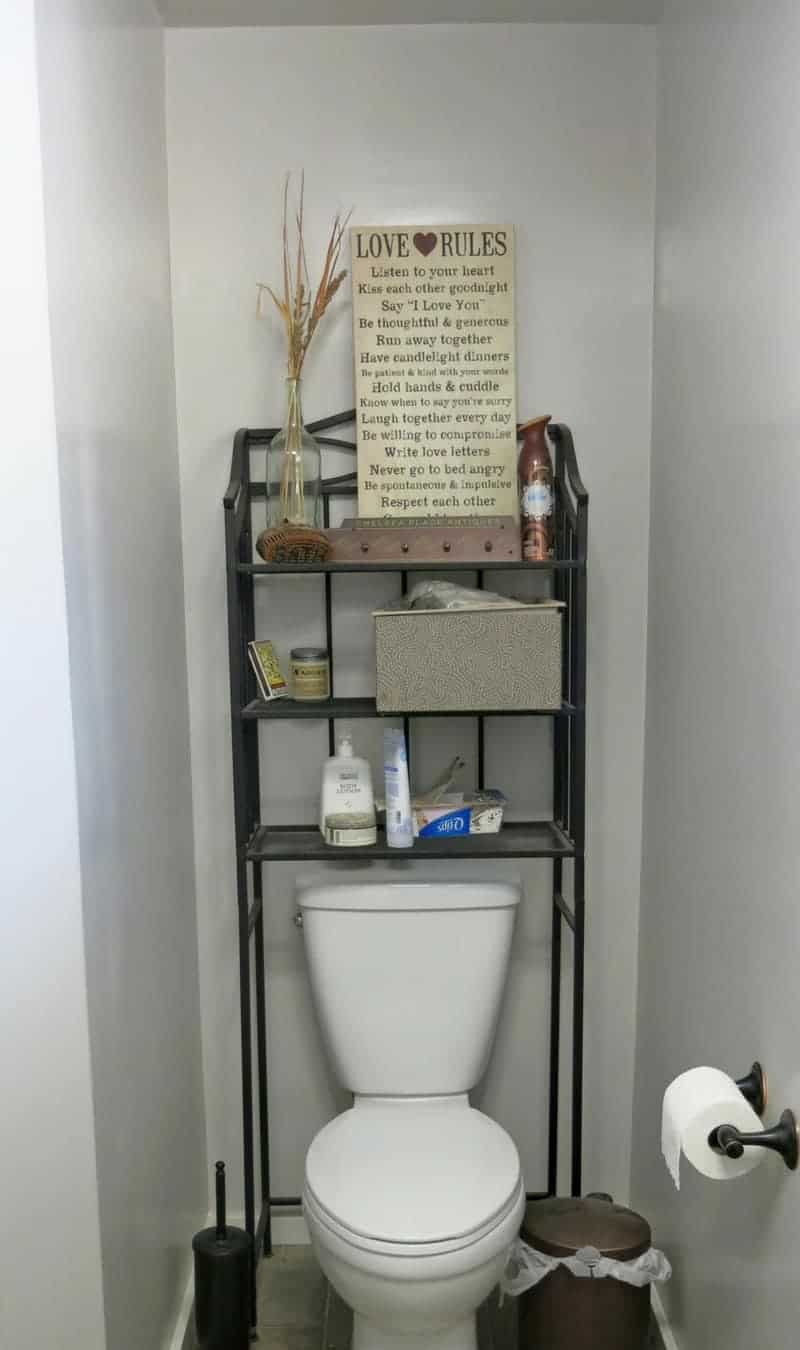 The boring bathroom toilet nook with bare white walls, and an over-the-toilet metal shelving unit cluttered with stuff.