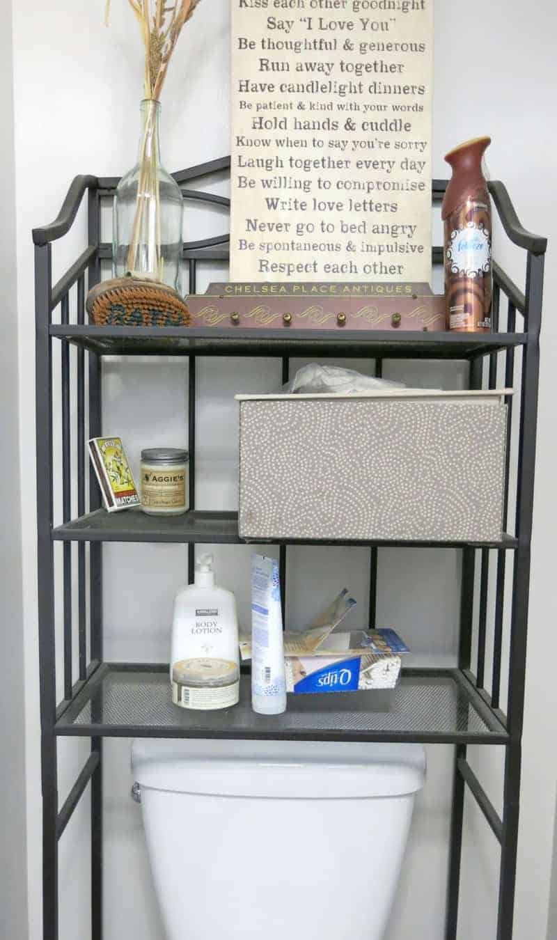 Cluttered black metal shelves above the toilet hold decorative signs, storage baskets, and toiletries.