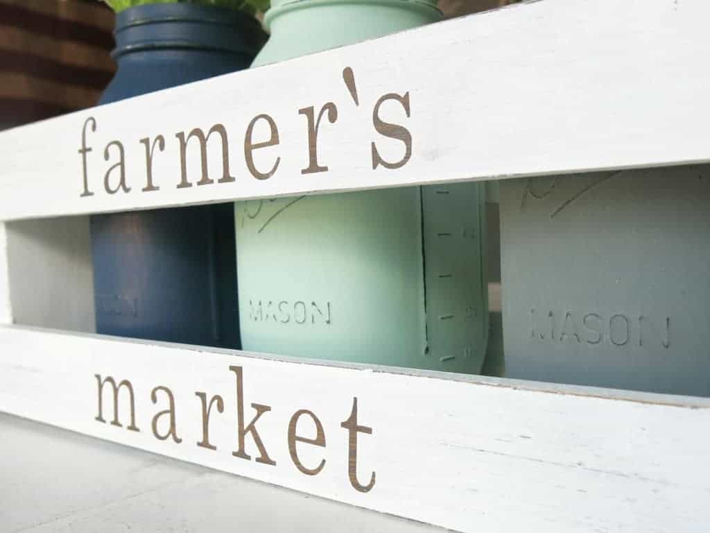 A close up side angled view of a white wooden crate with the words "farmer's market" on the side. The crate is filled with three painted mason jars, one blue, one mint green, and one gray.