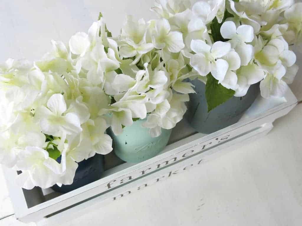 A rustic farmhouse centerpiece made from a wooden crate and painted mason jar vases with faux white flowers.