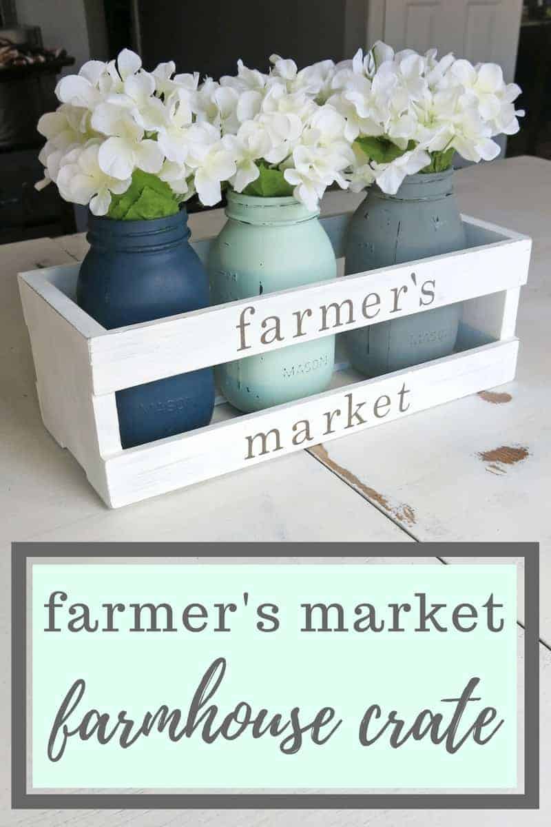 A look at the finished farmhouse crate centerpiece -- a white wooden crate filled with three painted mason jars, each holding a bunch of white flowers. Text across the bottom of the image says "farmer's market farmhouse crate"