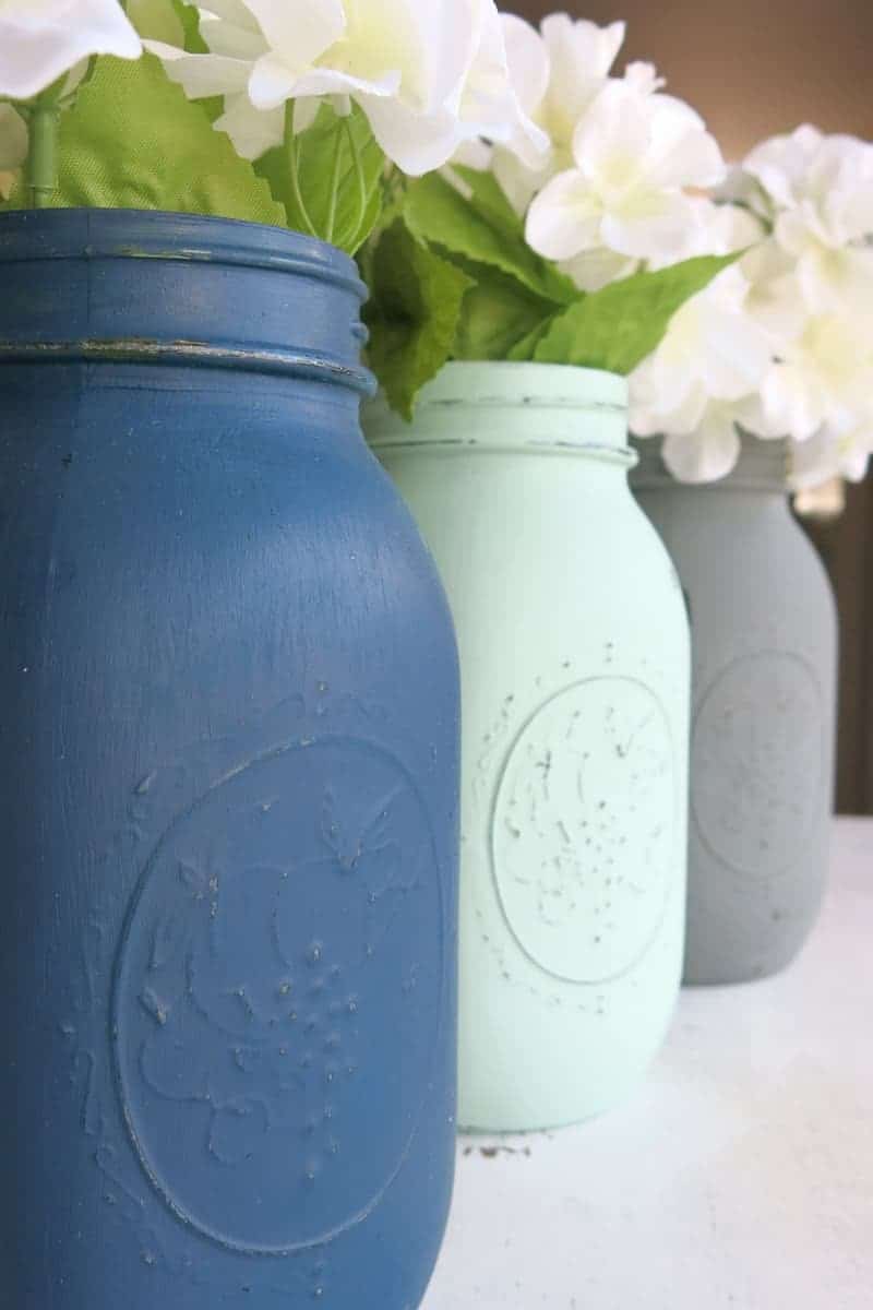 A close up look at three painted mason jars -- one blue, mint green, and gray -- with faux white flowers and leaves filling the jars.