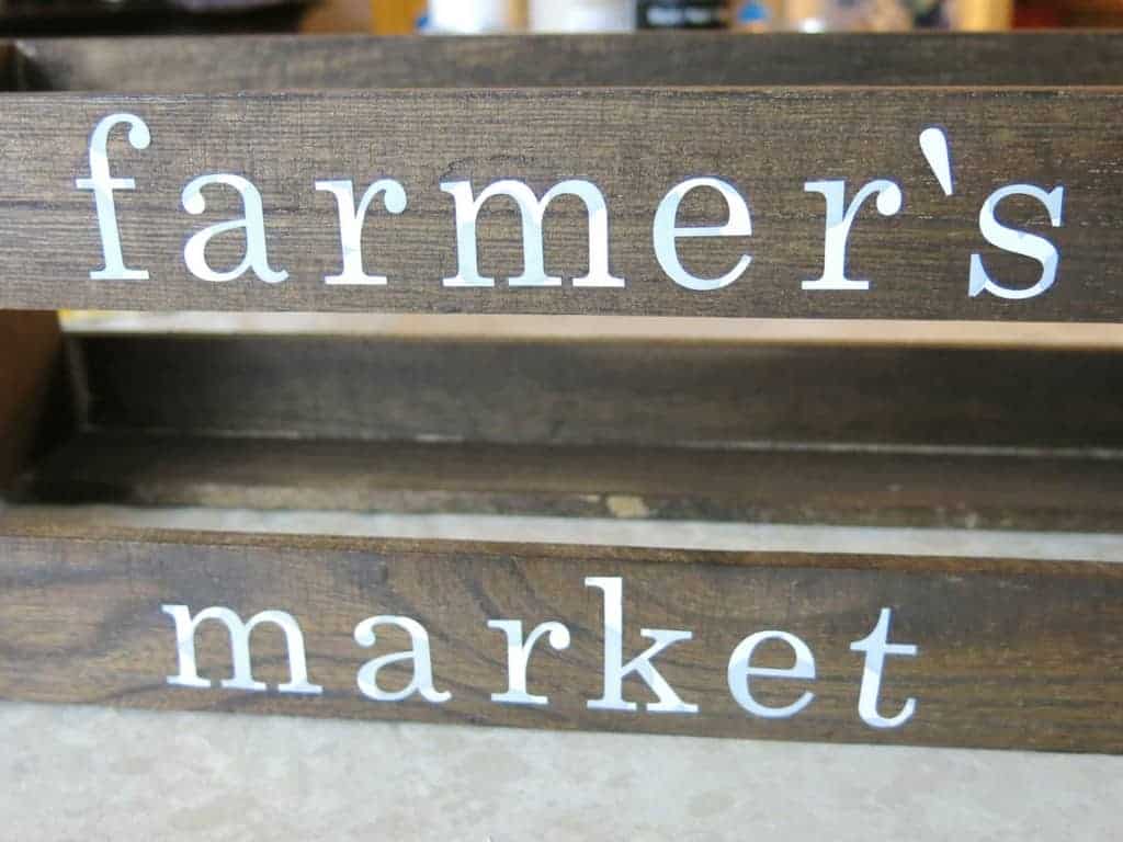 The words "farmer's market" spelled out with white vinyl letters applied to the side of a wooden crate.