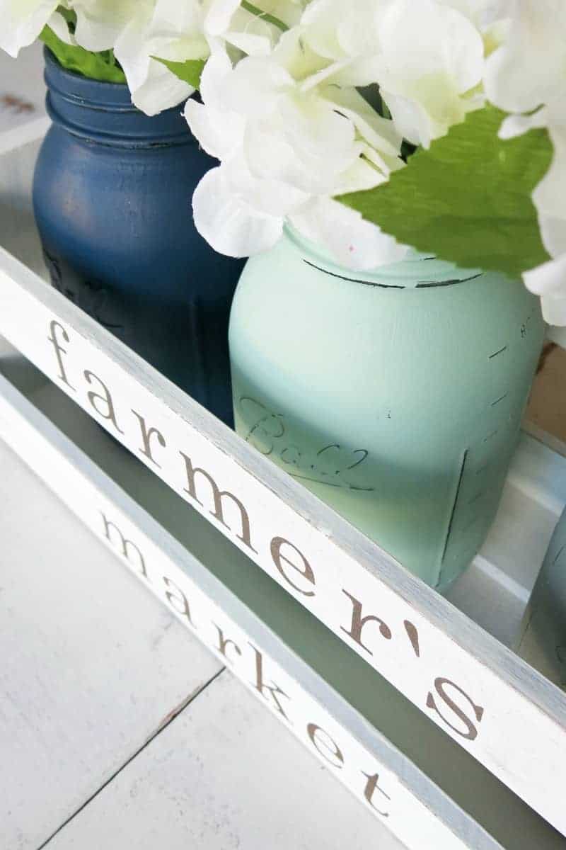 A close up image of two painted mason jar vases, each holding a bunch of white faux flowers. The mason jars sit in a white painted wooden crate with the words 