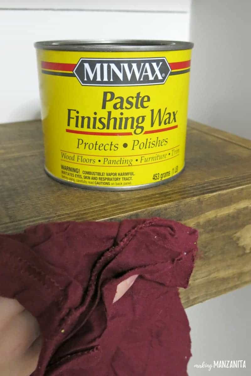 Can of Minwax Past Finishing Wax sitting on top of wooden floating shelf with hand applying the wax with a staining rag.