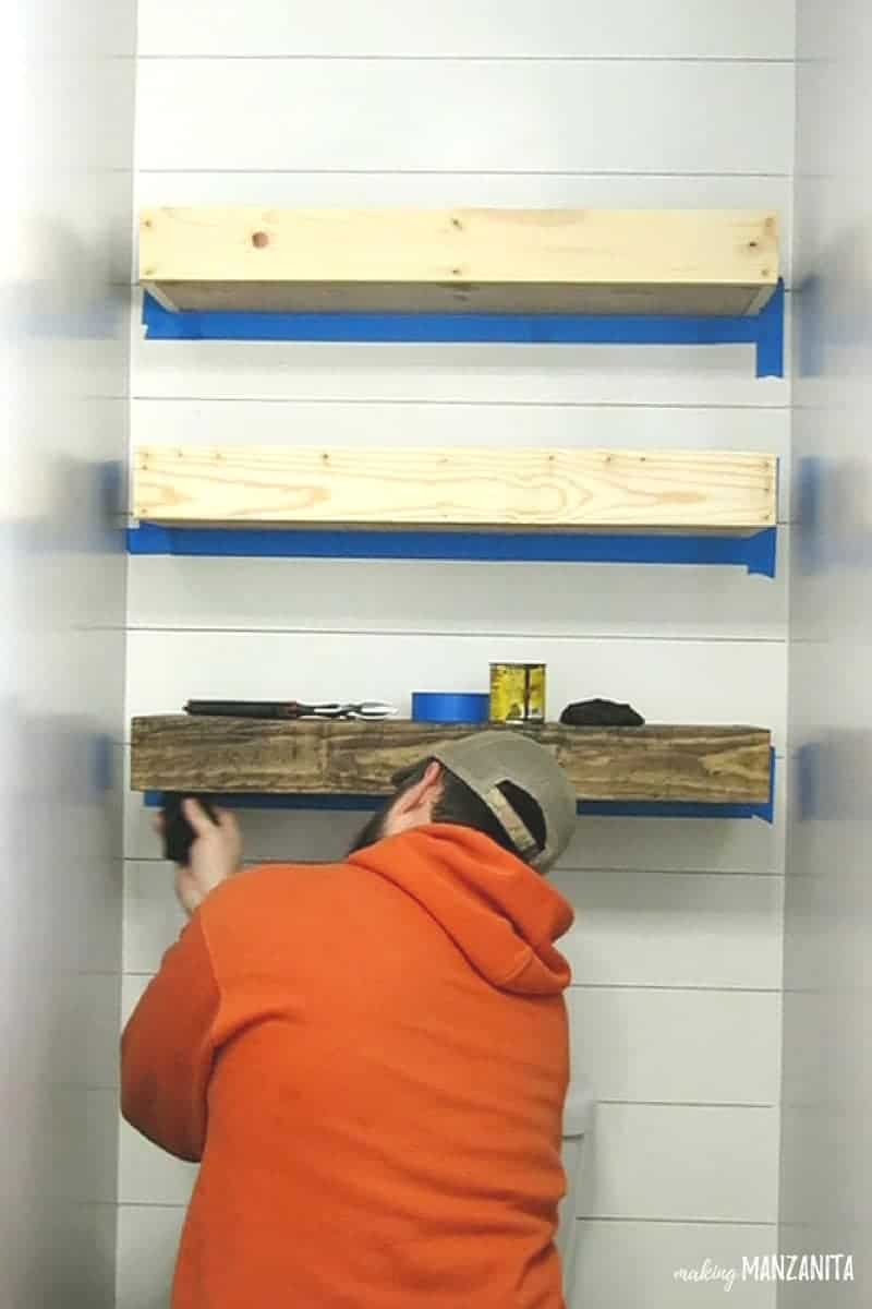 Man staining three floating shelves on wall with faux shiplap wall above toilet