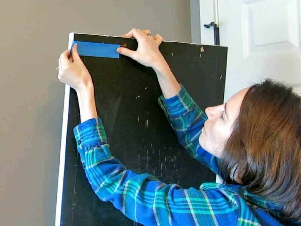 A women uses a piece of painters tape to measure the distance between the keyholes on the picture frame, and the top of the frame.