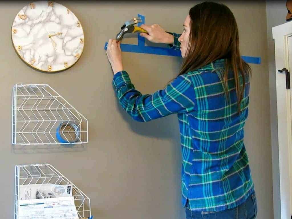 Using a hammer, put the nails in the wall using the keyhole guides marked on strips of painters tape. Using this method is the easiest way to hang pictures right on the first try!