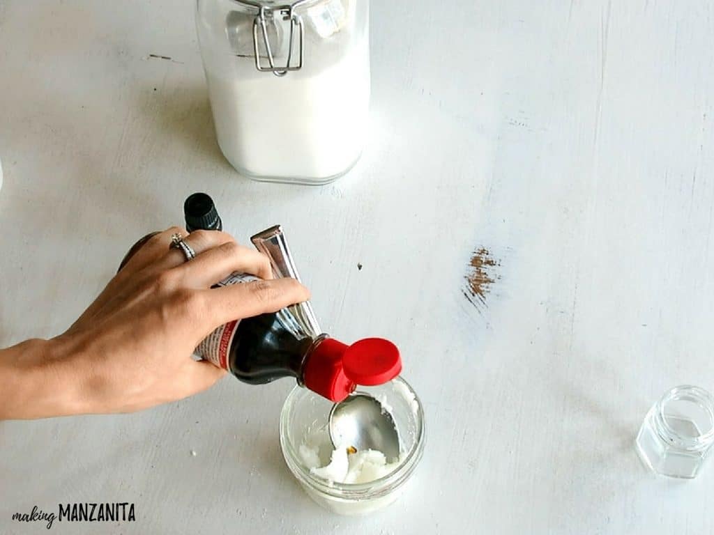 A woman adds a few drops of vanilla essential oil to a jar containing a sugar, coconut oil, and lavender essential oil lip scrub.