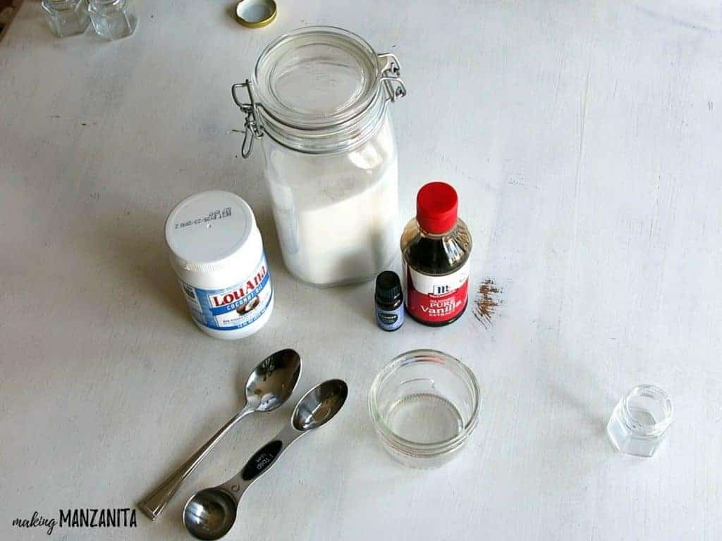 The supplies needed to make a DIY lavender vanilla lip scrub laid out on a table: coconut oil, sugar, lavender essential oil, vanilla extract, a jar, and spoons for mixing.