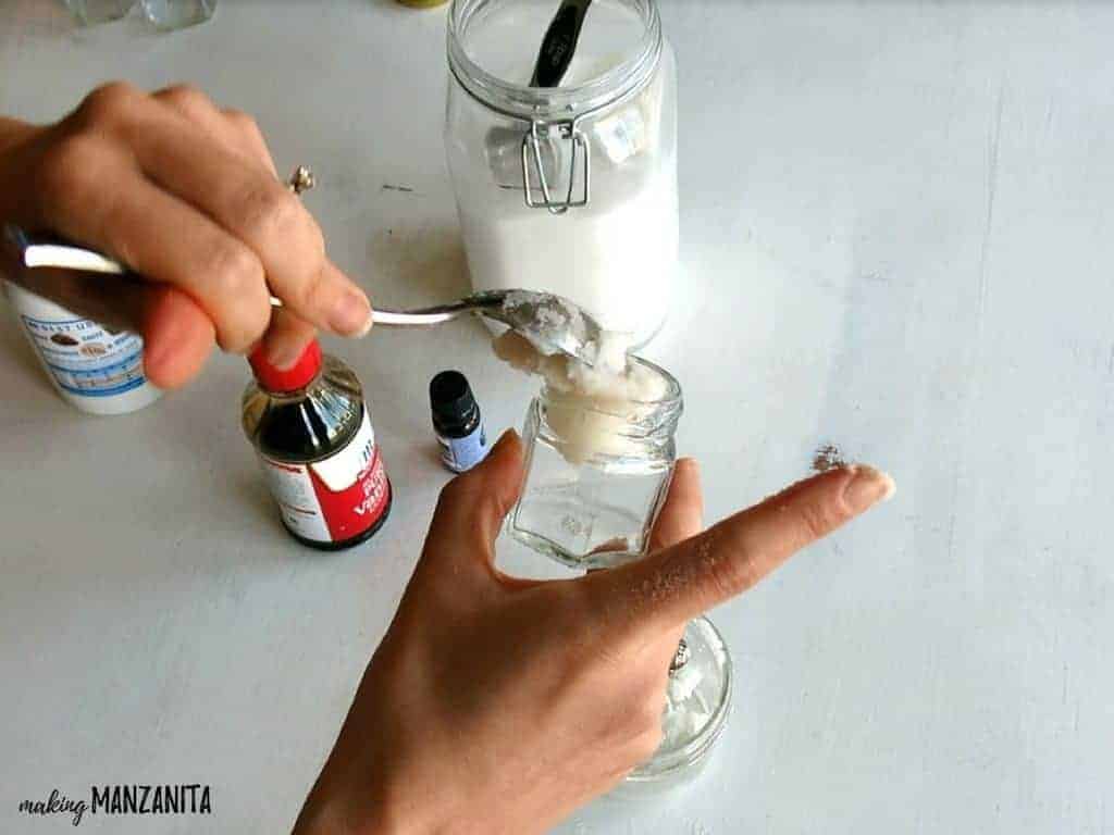 A woman spooning the lavender vanilla sugar scrub mixture into a small glass jar using a spoon.