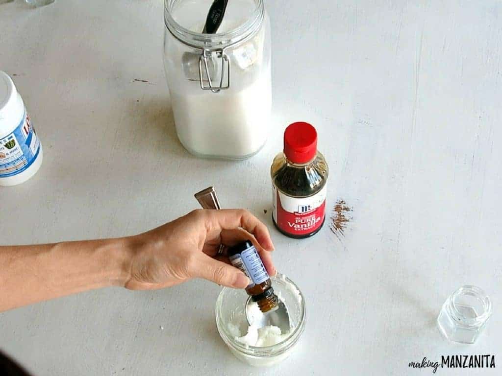 A woman adds drops of lavender essential oil to a jar with a sugar and coconut oil, creating a DIY lavender vanilla sugar lip scrub.