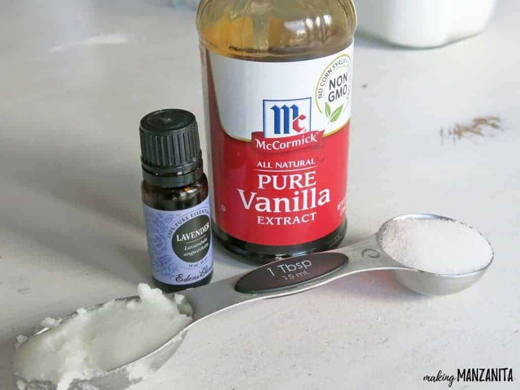 The supplies needed to make a DIY lavender vanilla lip scrub laid out on a table: coconut oil, sugar, lavender essential oil, vanilla extract, a jar, and spoons for mixing.