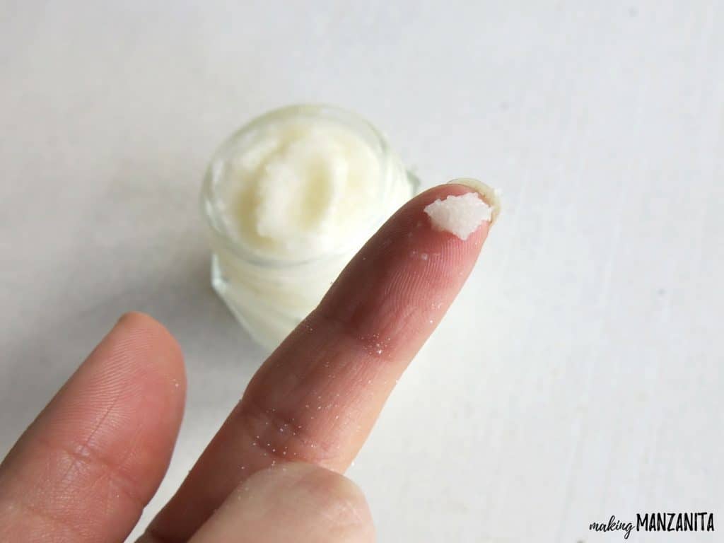 A small dollop of white lavender vanilla lip scrub sits on a woman's fingertip, the full jar visible in the background.