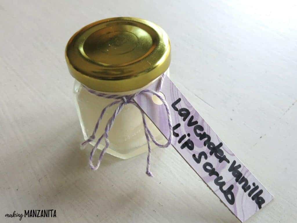 A small glass jar with a gold lid filled with a DIY lavender vanilla lip scrub, labeled with a purple gift tag.