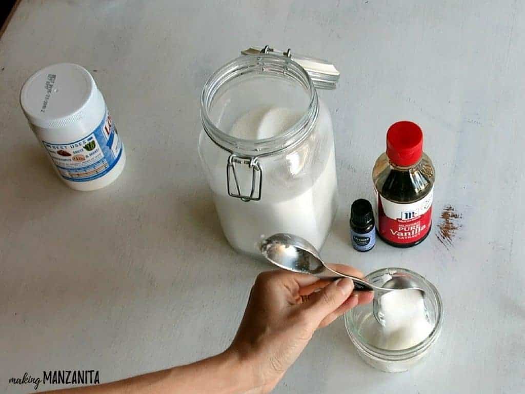 A woman pours a measuring spoon of sugar into a glass jar. Other lavender vanilla lip scrub ingredients sit on the table - coconut oil, vanilla extract, and lavender essential oil.