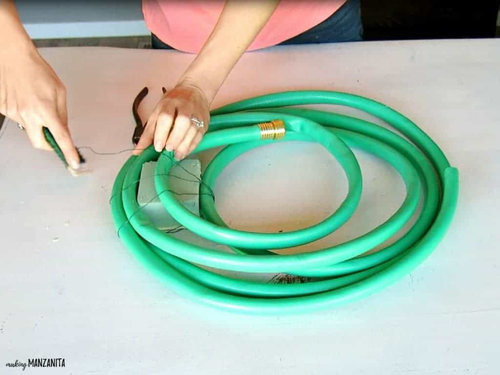 The first step in making a Spring garden hose wreath: a woman wraps a block of floral foam up in a green garden hose, and ties it together with floral wire. 