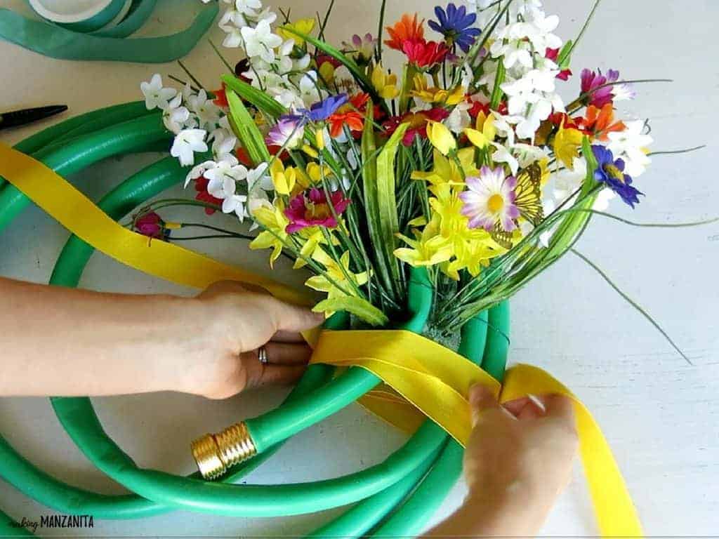 A woman wraps wide yellow ribbon around the floral foam and bunch of faux flowers, creating the floral feature of this upcycled garden hose wreath.